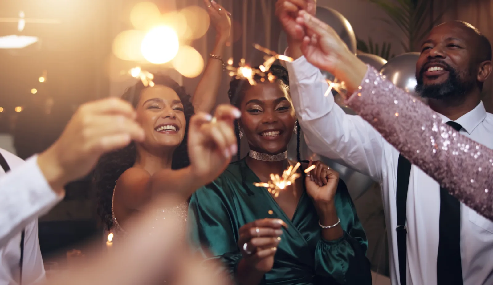 Smiling friends holding sparklers at a party with warm string lights and balloons behind them