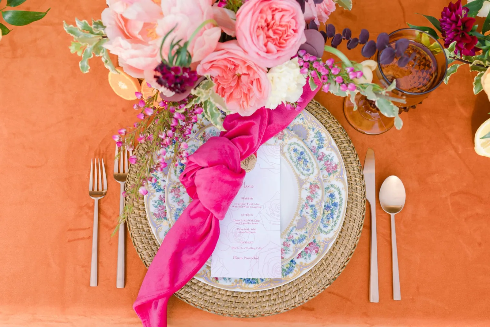 Overhead view of a table set with floral china, a pink velvet napkin, a printed menu card and pink roses on a peach tablecloth